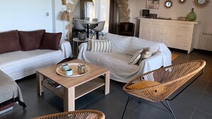 A modern living room with a sofa, a coffee table, and a wicker chair. There is a dining area with a table and chairs, and a kitchen area with cabinets and a countertop. - Villa Pals very beautiful sea view and Medes islands, private swimming pool (Pals - Masos de Pals)