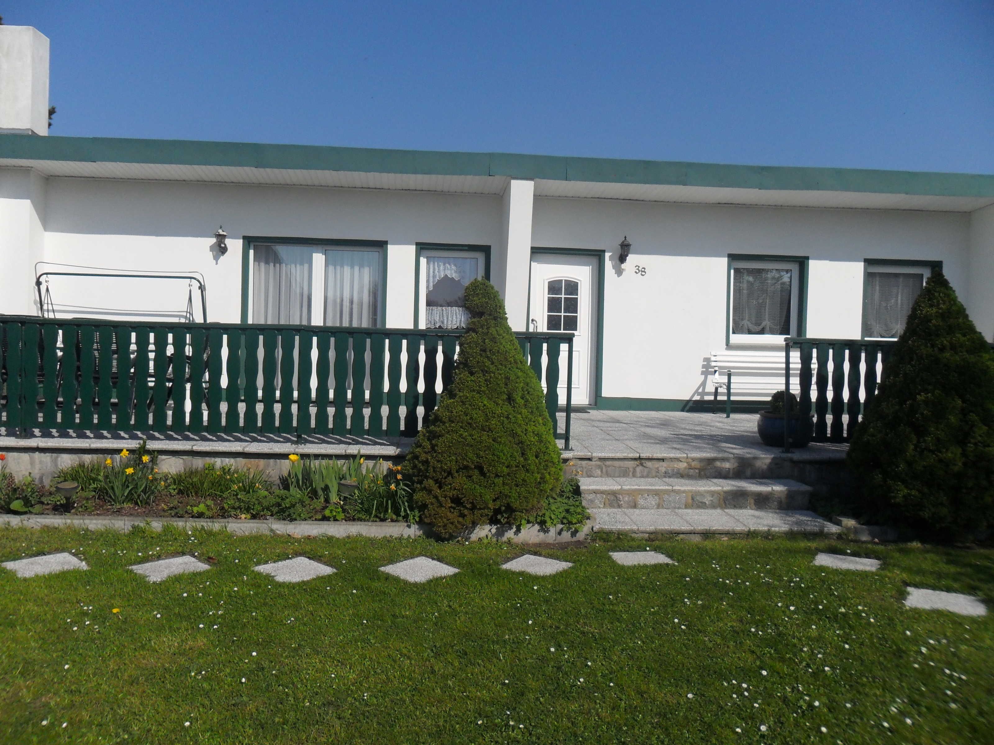 A building with a green roof, white walls, and a number '38' on the door. There is a bench and a small table outside.