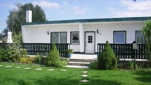 A single-story building with a white exterior and a green roof. There are two doors, a small porch with a black railing, and a small garden with trimmed bushes and a lawn.