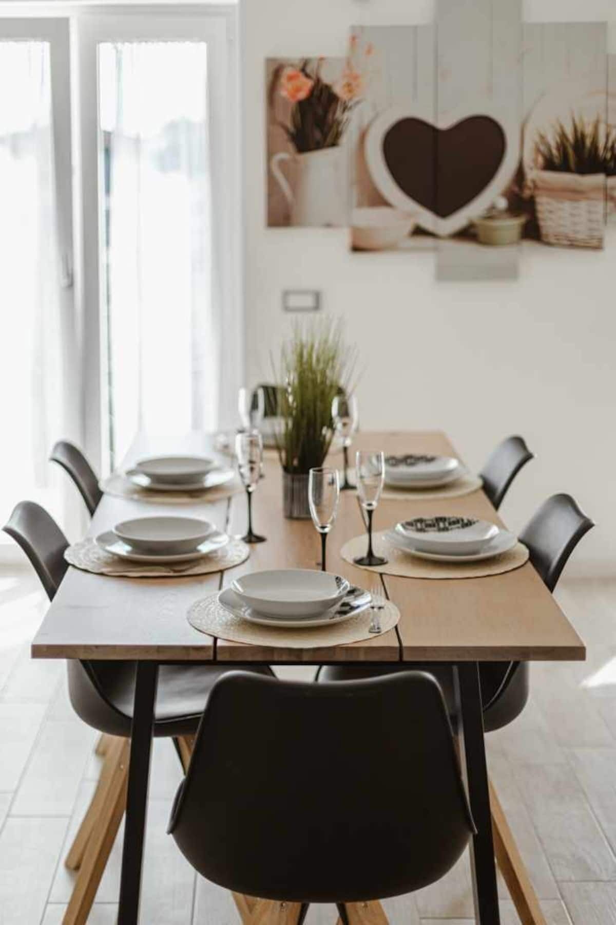 Une table à manger dressée avec des assiettes, des verres et des couverts, entourée de chaises noires dans une pièce lumineuse avec un mur blanc et une décoration en forme de cœur.