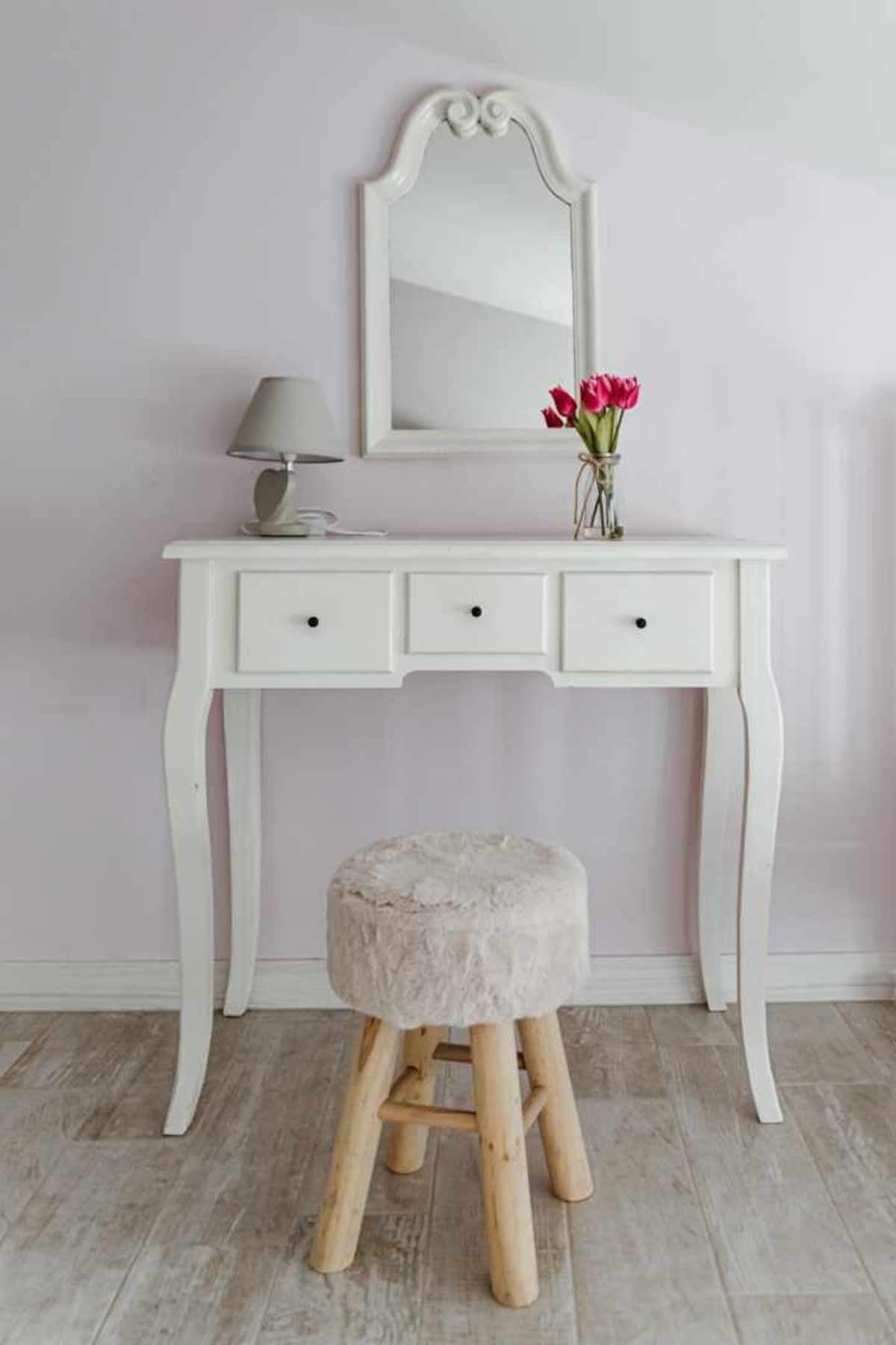 A white vanity with a mirror, three drawers, a lamp, and a stool with a vase of flowers.