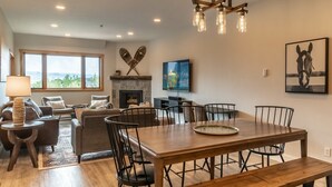 A spacious living area with a large wooden dining table, comfortable seating, a fireplace, and a scenic view through large windows.