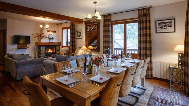 Un coin repas avec une table en bois dressée pour six personnes, une cheminée et une vue sur l’extérieur grâce à de grandes fenêtres.