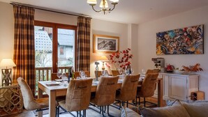 Un coin repas avec une table dressée pour six personnes, des chaises, un lustre, un tableau et une vue sur un bâtiment à l’extérieur.