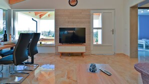 A modern living room with a flat-screen TV on a wooden stand, a dining area with chairs and a table, and a large window offering a view of the outdoors. - House 2065/32772 (Istria - Žminj), 20000m from the beach (Žminj)