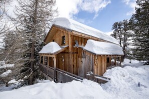 Une cabane en bois, dont le toit est recouvert de neige, entourée de neige et d’arbres.
