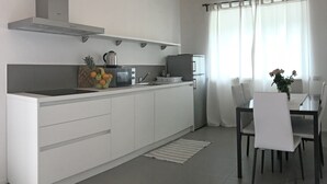 Ett modernt kök med vita skåp, en fläktkåpa i rostfritt stål, en mikrovågsugn och ett kylskåp. Det finns en matplats med ett bord och stolar, och ett fönster med tunna, genomskinliga gardiner.