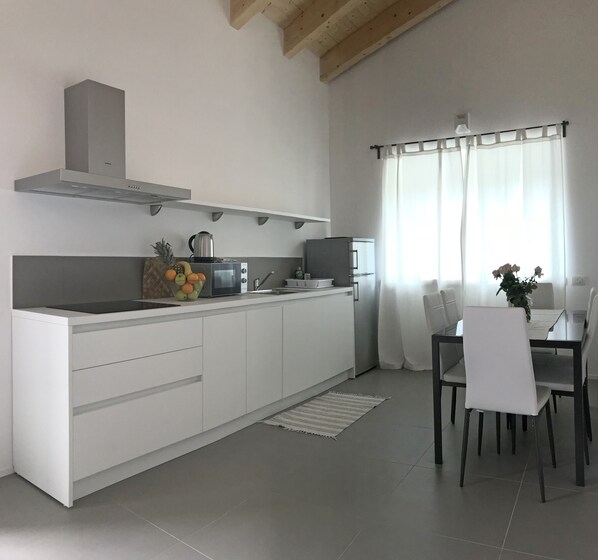 Een moderne keuken met witte kasten, een roestvrijstalen afzuigkap, een magnetron en een koelkast. Er is een eethoek met een tafel en stoelen, en een raam met dunne gordijnen.
