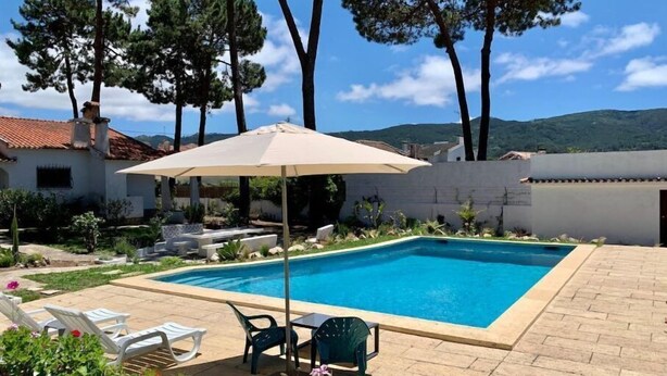 Et rektangulært swimmingpool med flisekant, omgivet af en terrasse med stole og en parasol.