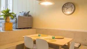 Un coin repas avec une table en bois et des chaises blanches, une horloge au mur et une plante sur le rebord de la fenêtre.
