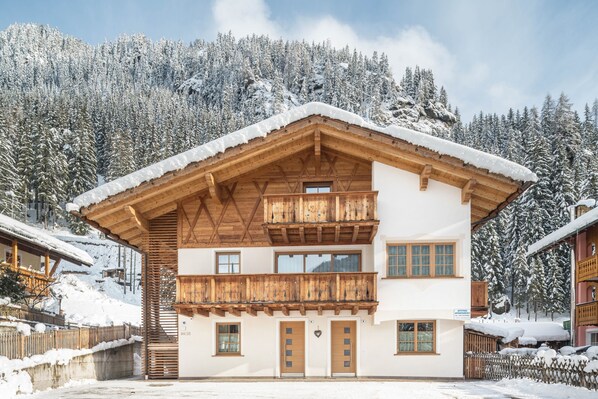 A two-story wooden chalet with a balcony, set against a snowy mountain backdrop.