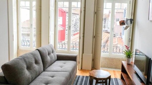 Una sala de estar con un sofá gris, una mesita redonda y un televisor sobre un mueble de madera. Amplias ventanas con cortinas ofrecen vistas a un edificio rojo y a un balcón.