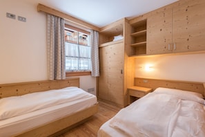 A room with two beds, wooden wardrobe, a window with curtains, and a small bedside table.