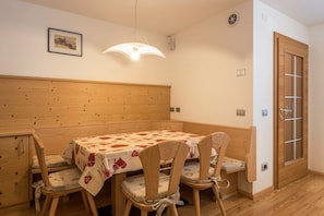 A dining area with a table covered in a patterned tablecloth, wooden chairs, a wooden bench, a pendant light, a framed picture on the wall, and a door with a glass panel.