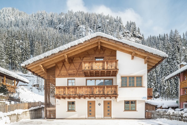 A two-story wooden chalet with a balcony, set against a snowy mountain backdrop.