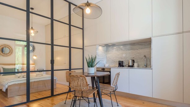 A modern kitchen with a marble backsplash, a dining area with wicker chairs, and a bedroom with a large window.