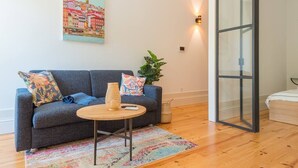 A modern living room with a grey sofa, a round wooden coffee table, and a colorful rug. There is a painting of a cityscape on the wall, a plant, and a sliding glass door.