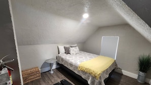 A small, sloped-roofed bedroom with a bed, bedside table, and a potted plant. - (PPV1) The Attic | Historic Hurricane (Hurricane)