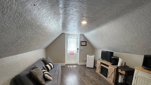 A small, cozy room with a sloped ceiling, a gray sofa with striped cushions, a flat-screen TV, a small cabinet, and a white door with an exit sign. - (PPV1) The Attic | Historic Hurricane (Hurricane)