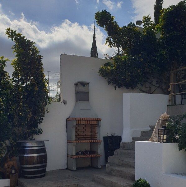 Un patio con un horno de ladrillo, un barril grande y una pared blanca con una ventana pequeña.