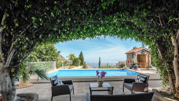 A poolside area with wicker furniture, a table with flowers, and a view of a house and landscape. - Vacation Home 'Casa Reina Del Atlántico' with Sea View, Private Pool and Wi-Fi (Candelaria)