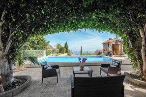 Un espacio junto a la piscina con mobiliario de mimbre, una mesa con flores y vistas a una casa y un paisaje.