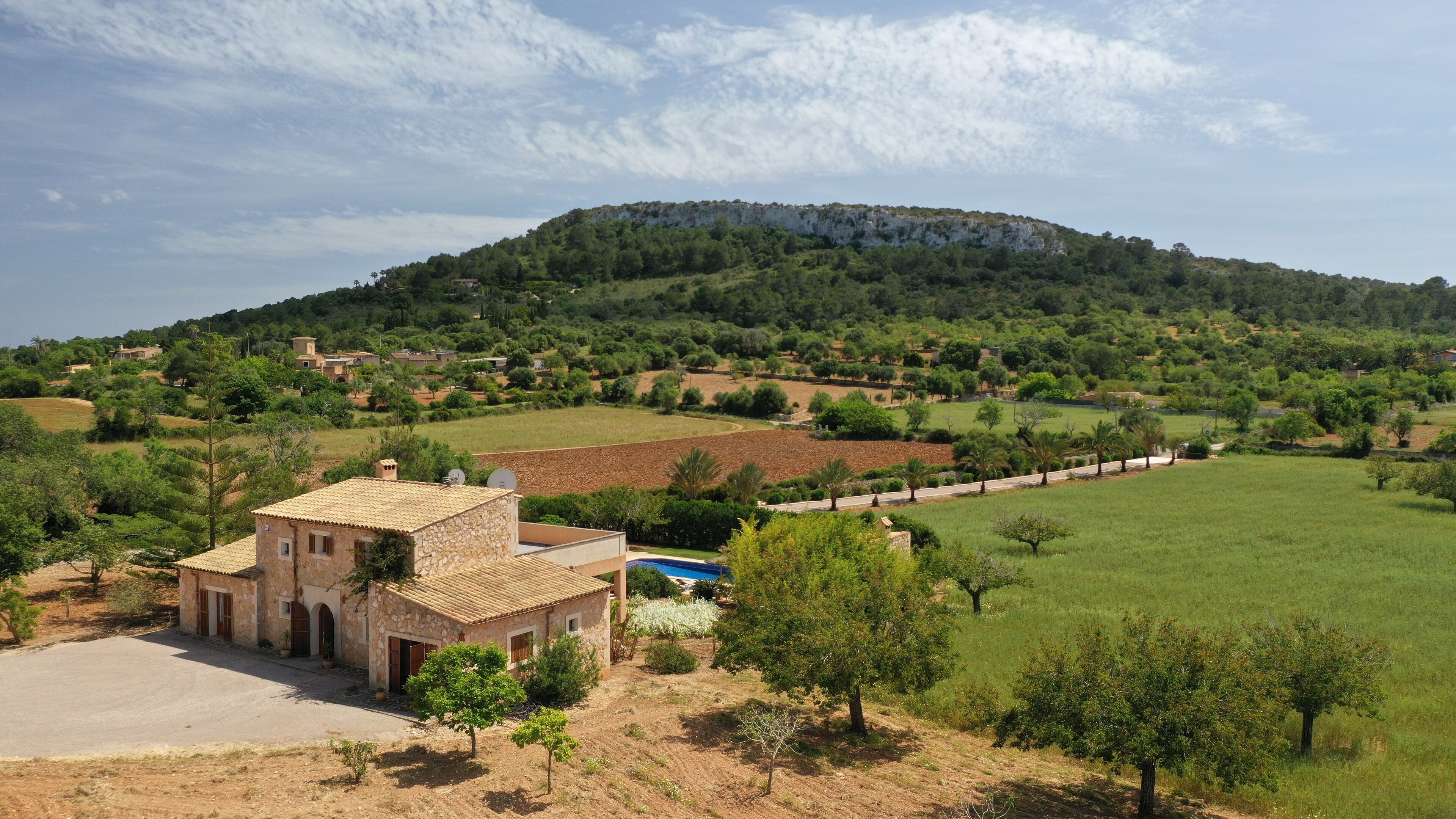 A stone building with a tiled roof, a swimming pool, and a view of a green valley with a hill in the background.