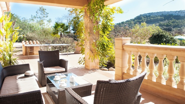 An outdoor patio with wicker furniture, a table set with a teapot and cups, and a view of the surrounding landscape including trees and hills. - Country house with pool and beautiful views (Cas Concos)