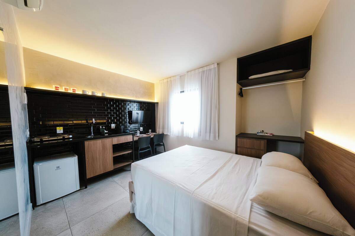 A hotel room with a bed, a desk with a chair, a kitchenette, and a TV.
