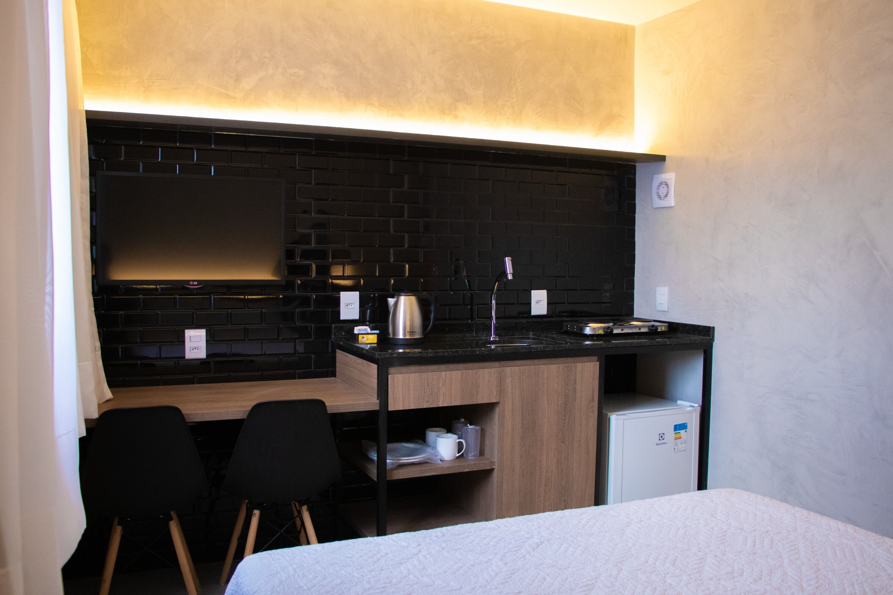A compact hotel room with a kitchenette, two black chairs, a bed, and a small refrigerator.