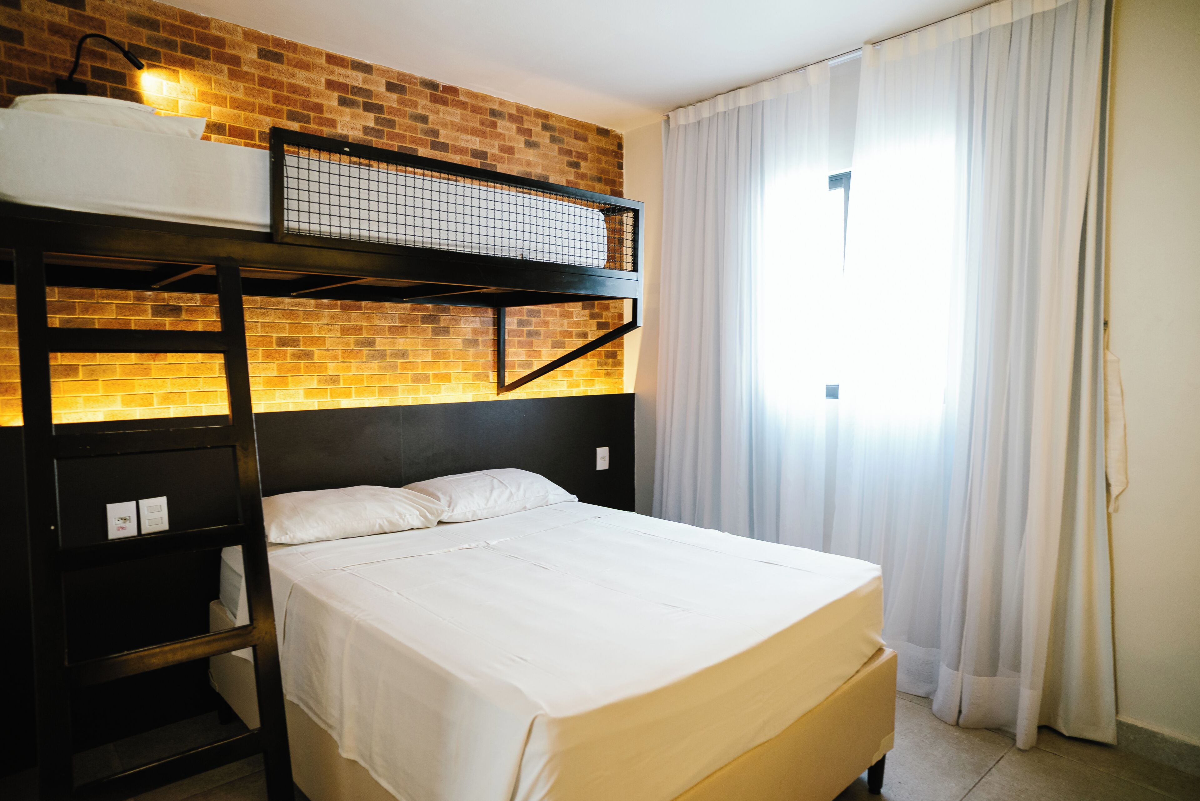 A bunk bed with a headboard against a brick wall, a window with curtains, and a bedside table.