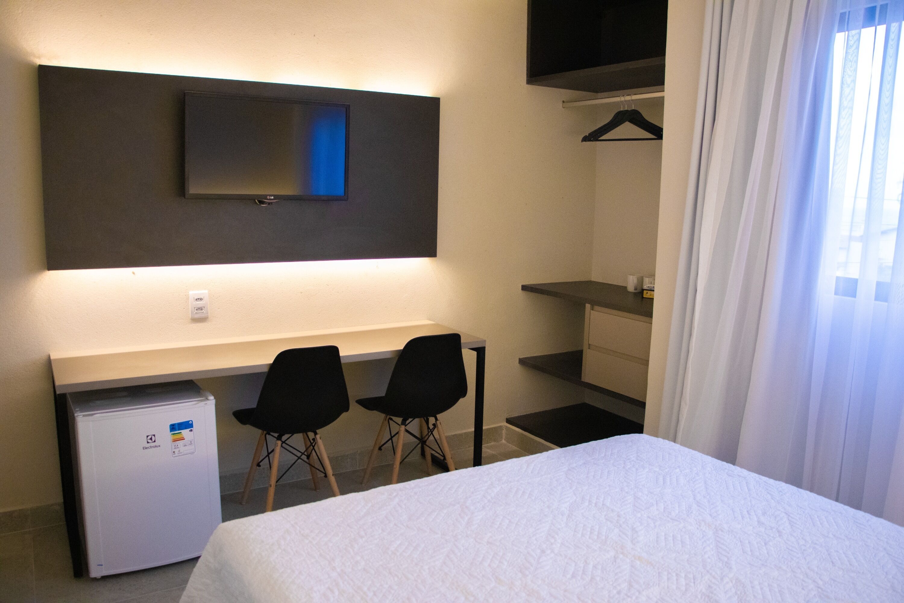 A hotel room with a bed, a desk with two chairs, a television mounted on the wall, a small refrigerator, and a window with curtains.