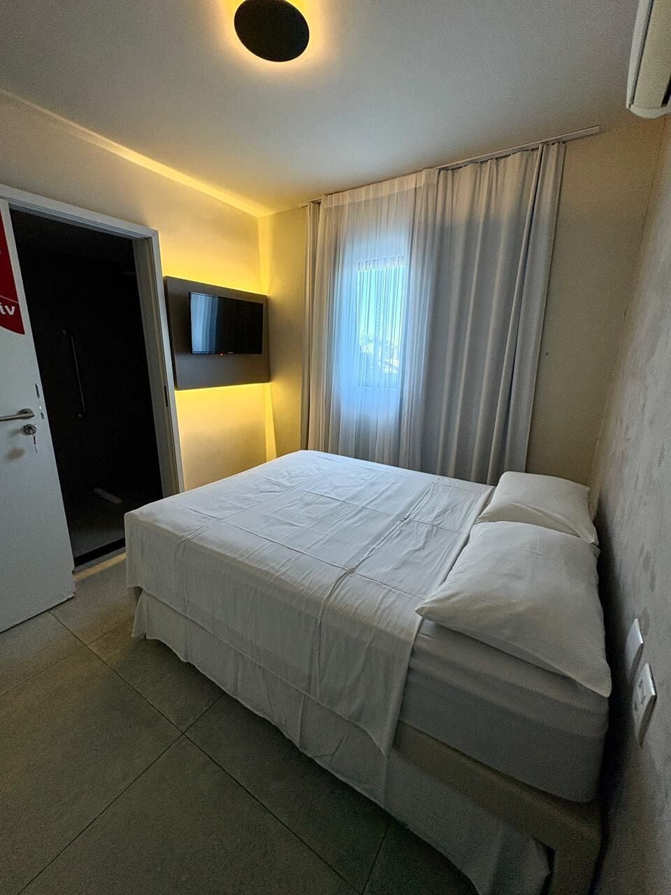 Habitación de hotel con una cama, un televisor montado en la pared, una puerta y una ventana con cortinas.