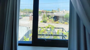 A room with a large window, curtains, and a view of a parking area and greenery outside. - Liiv Costeira - Natal Ponta Negra (Natal)