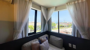A room with a large window, white curtains, a dark-colored sofa with cushions, and an air conditioning unit on the wall. - Liiv Costeira - Natal Ponta Negra (Natal)