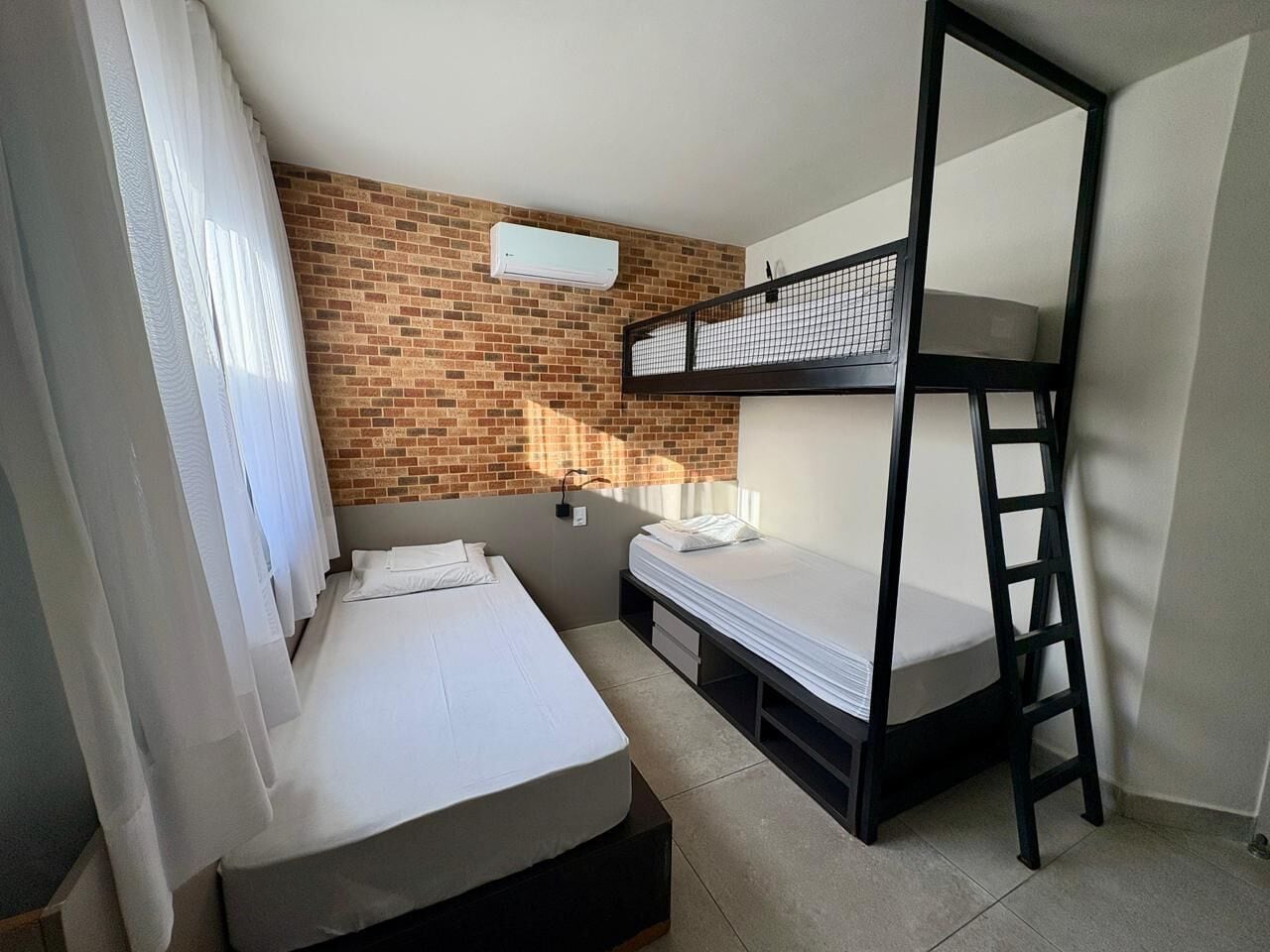 A room with a bunk bed, a single bed, a brick wall, a window with curtains, and a white air conditioning unit.