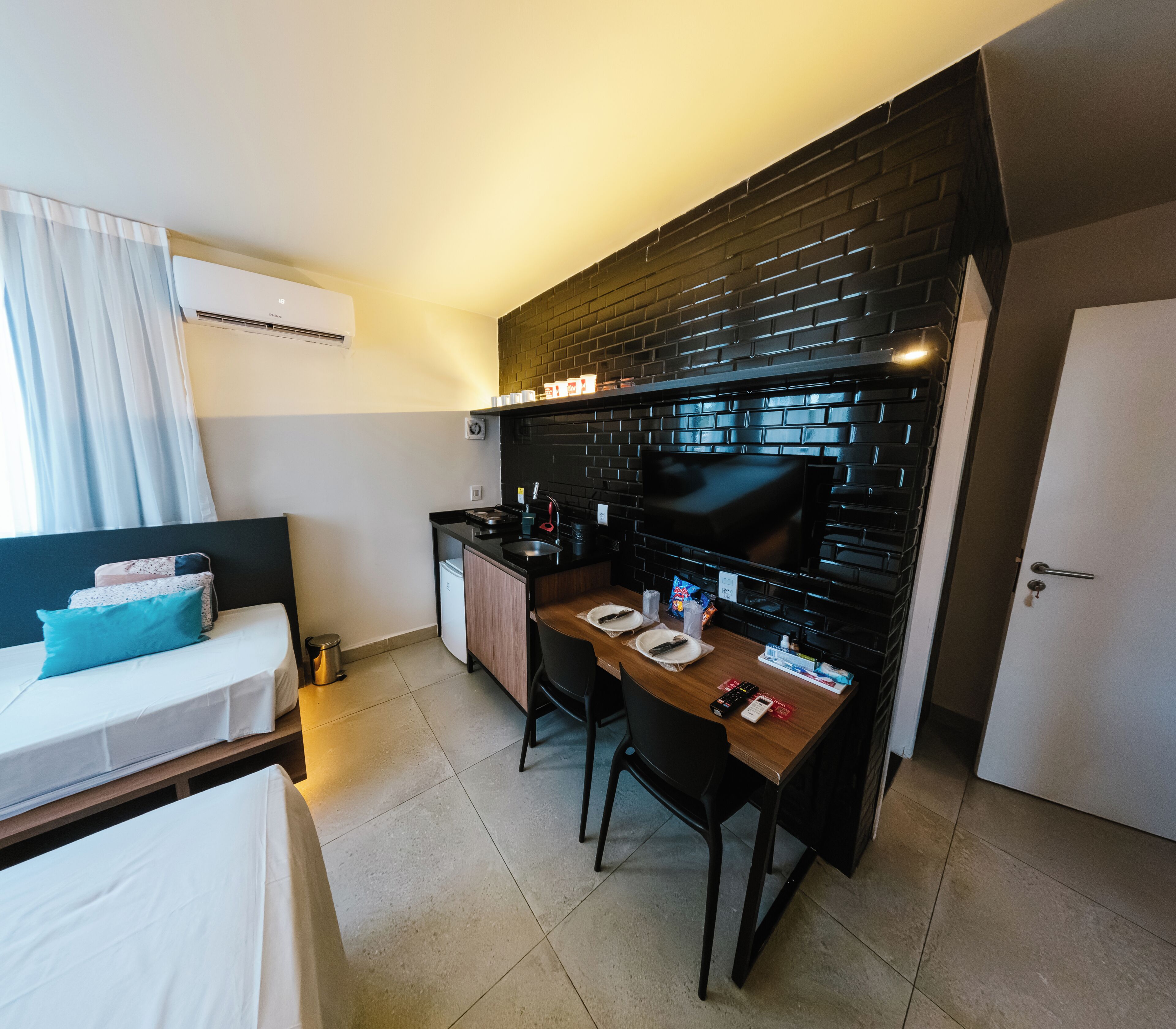 Habitación de hotel compacta con kitchenette, una cama con almohada azul, una mesita con televisor y una puerta blanca.
