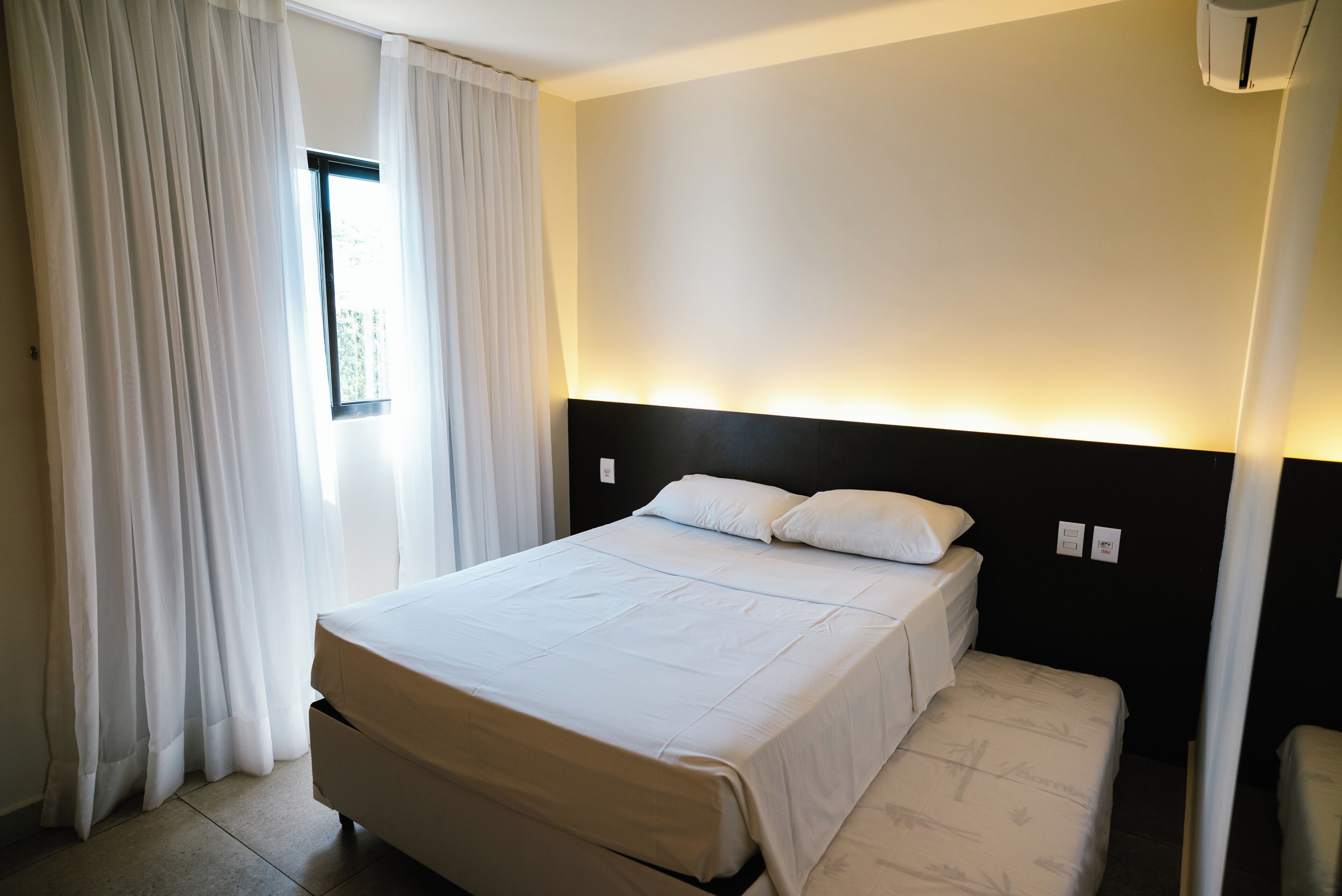 A hotel room with a bed, curtains, a window, and a wall-mounted air conditioning unit.