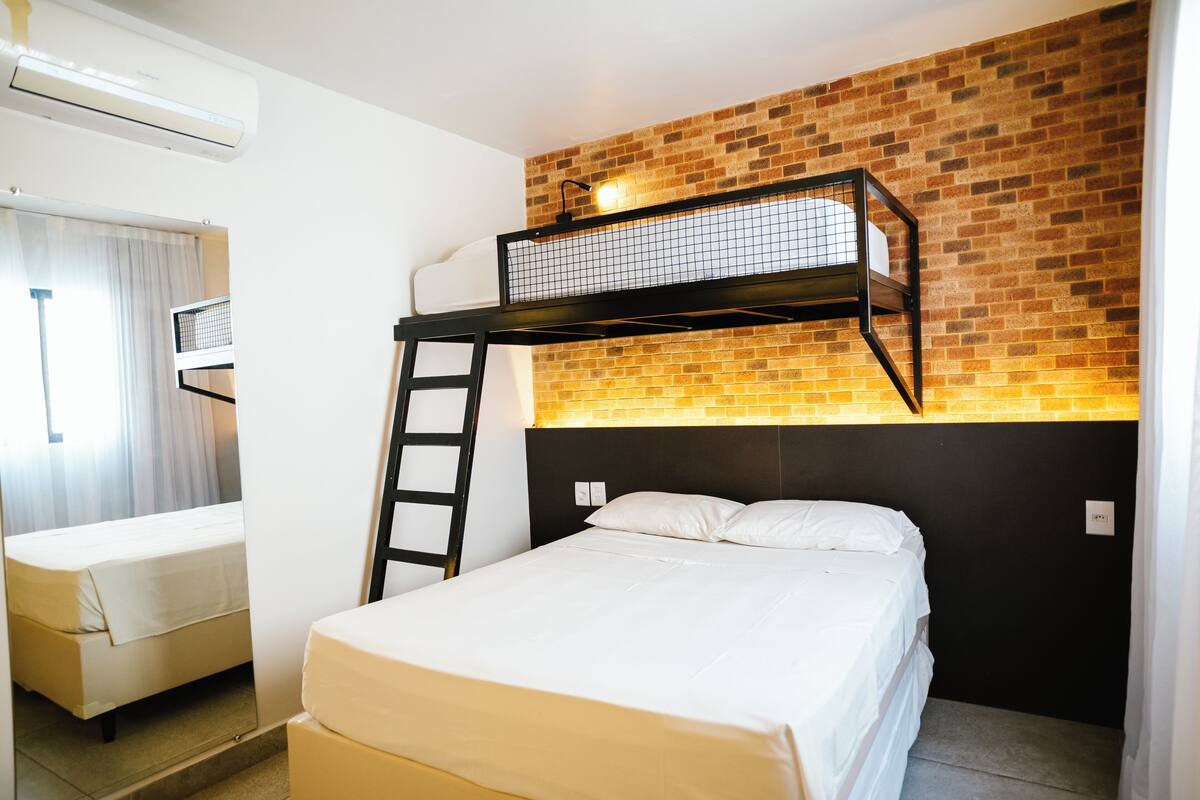 A bunk bed with a ladder, a single bed, a window with curtains, and a brick wall.