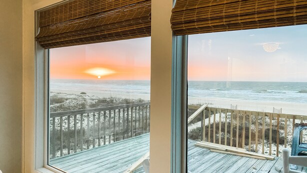 Een kamer met een groot raam dat uitkijkt op een strand, een houten terras en een uitzicht op de oceaan tijdens de zonsondergang.