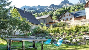 A rustic outdoor seating area with a table and chairs, surrounded by greenery and stone walls, with mountain views in the background. - Private terrace, warm & cosy lounge with fireplace (Vaujany)