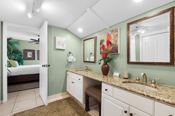 Baño con lavamanos doble, tocador y un espejo grande. Se ve una cama en la habitación contigua.