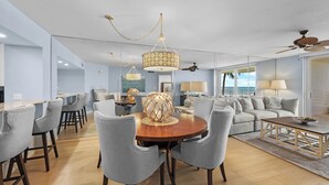 Un salón moderno con una mesa redonda de madera, sillas tapizadas en gris, un ventilador de techo y un ventanal con vistas al exterior.