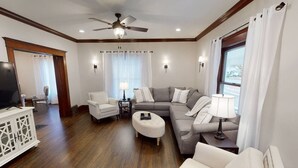 Un salón amplio con un sofá gris, sillones blancos, una mesa de centro y un televisor sobre un mueble de madera.