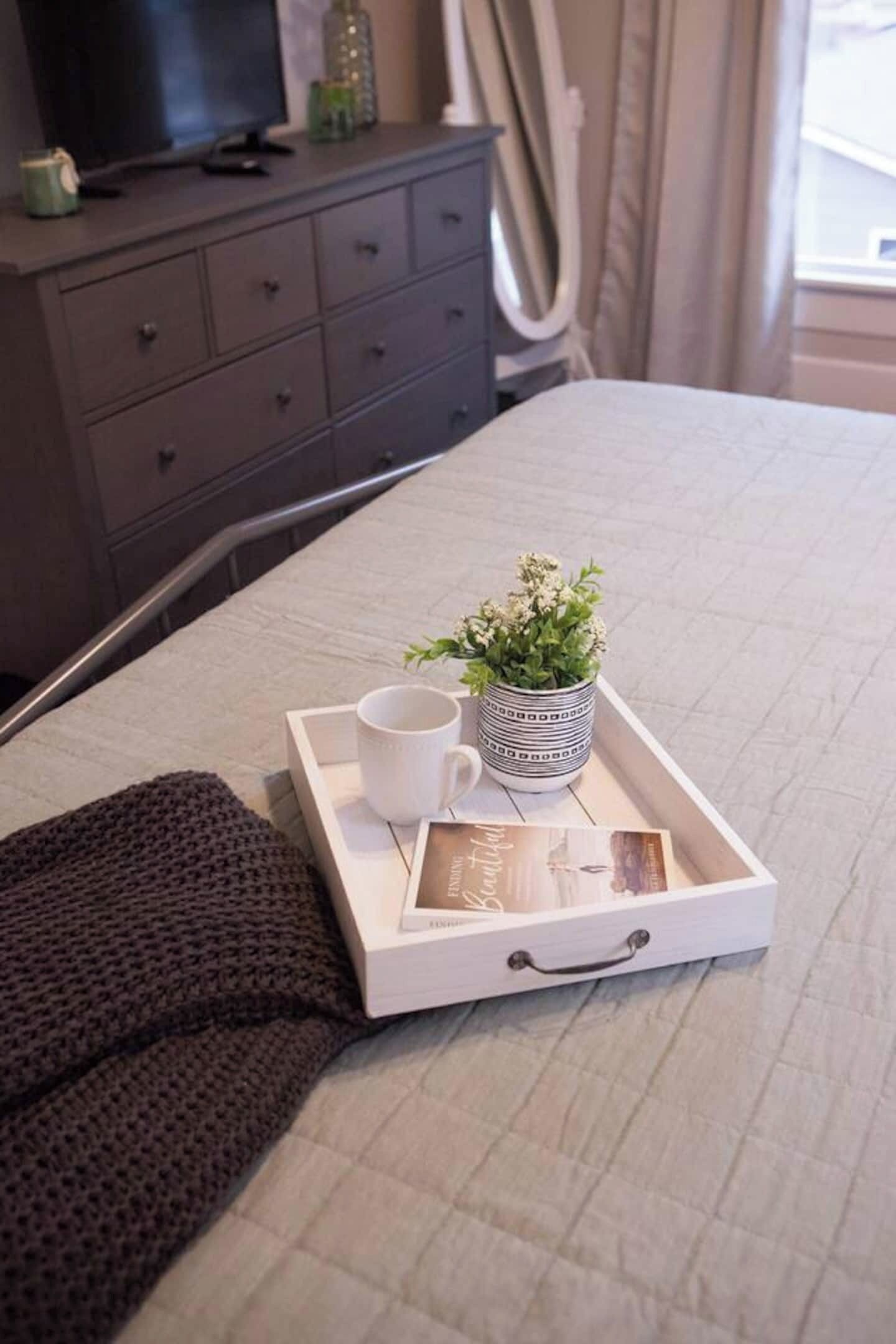 A neatly made bed with a tray containing a mug, a small potted plant, and a framed picture.