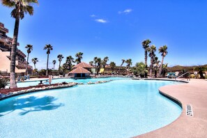 A large swimming pool with palm trees and a small building in the center. - Seaside Serenity @ MP206 | Surf and Pools (Port Aransas)