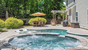 A hot tub in a backyard with a patio table and chairs, surrounded by trees and a wooden fence. - Pleasant Place New Build + Pool Great for Families (Mount Pleasant)