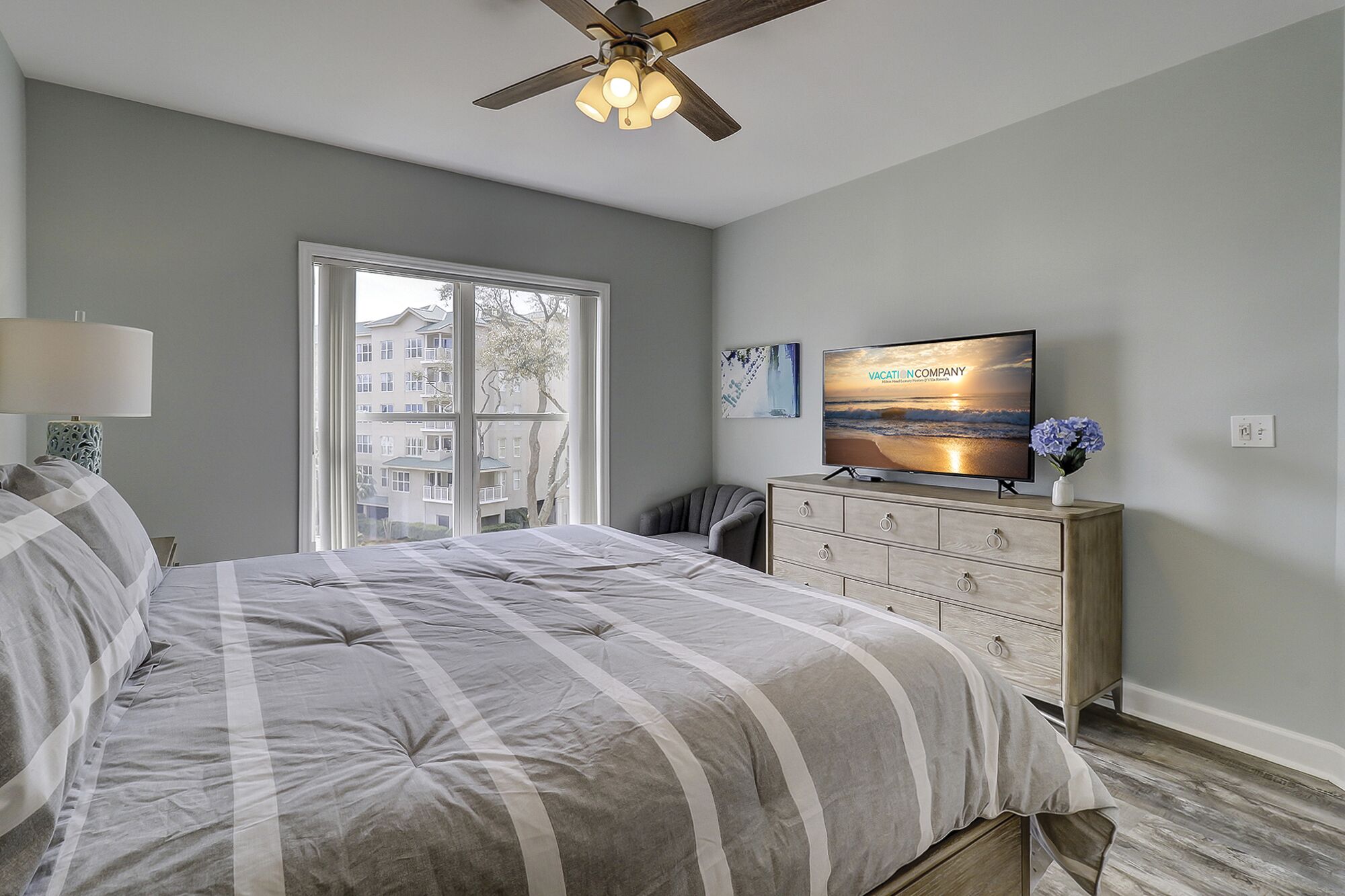 A bedroom with a bed, a dresser, a television displaying 'VACATIONCOMPANY', and a window with a view of buildings and trees.