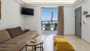 A modern living room with a sofa, a coffee table, and a TV. There is a door leading to a balcony with a pool and chairs. - Beachfront Villa Alexandra (Σκάλα)