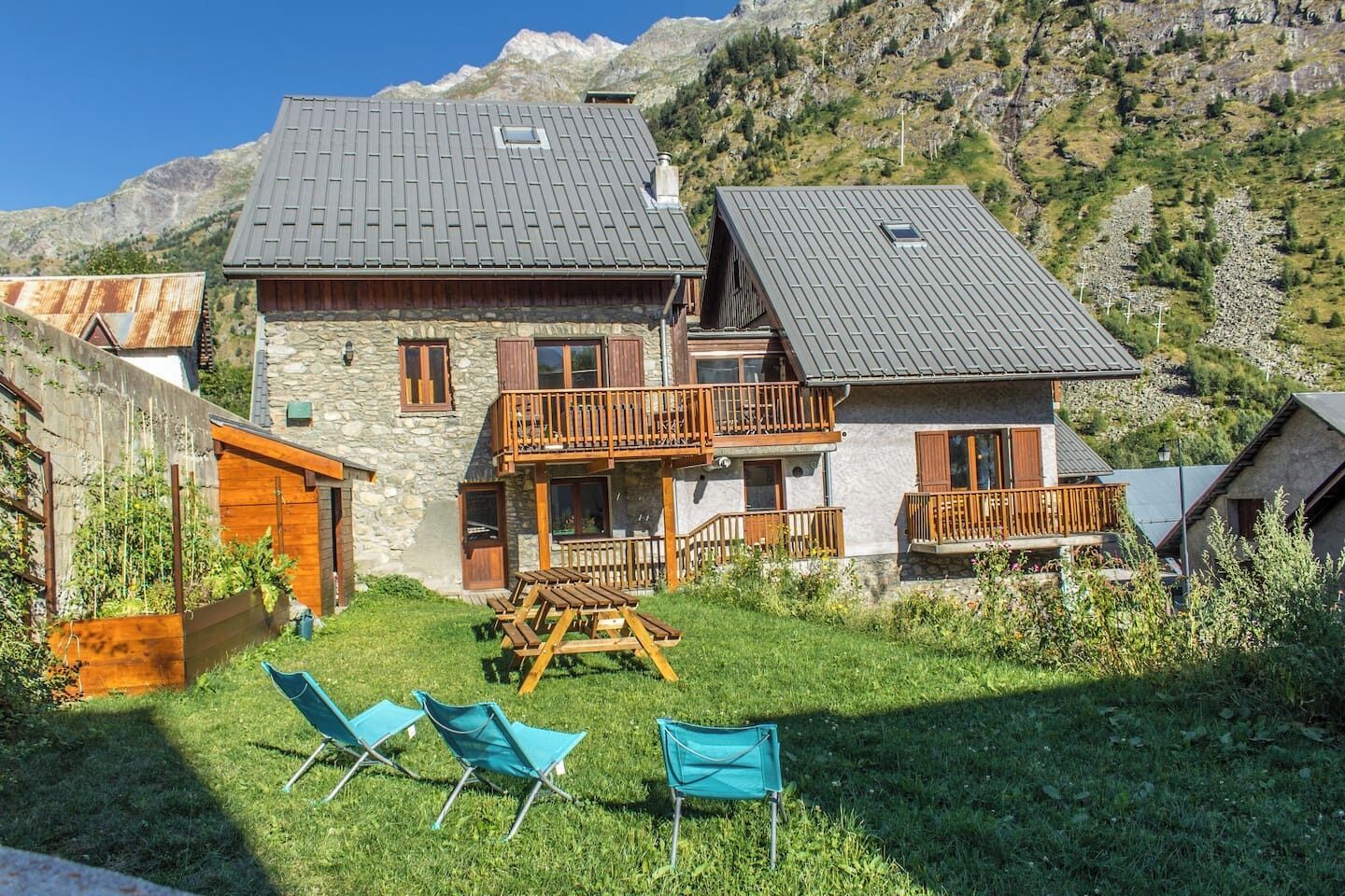 A two-story stone house with a balcony, a wooden deck, and a grassy yard with two blue chairs.